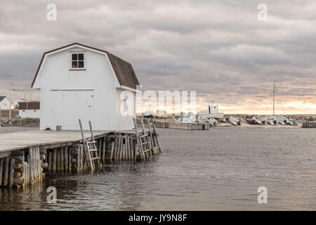 White boathouse on dock on a cloudy day at sunset near Twillingate, Newfoundland, Canada. Fishing boats lined up in the distance on shore. Light rippl Stock Photo