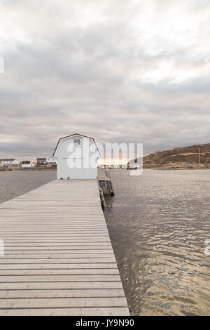 White boathouse on long dock on a cloudy day at sunset near Twillingate, Newfoundland, Canada. Houses in distance. Light ripples in water. Stock Photo