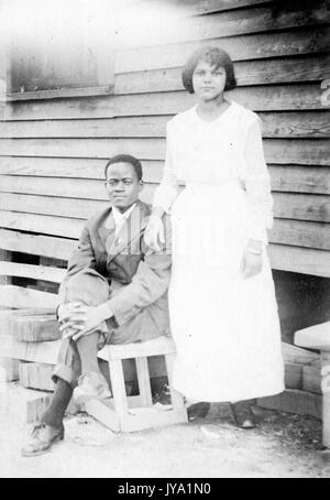 African-American couple posing in front of a house with wood siding, wife wearing white dress and standing, husband sitting on a wooden crate with his hand on his knee, 1910. Stock Photo