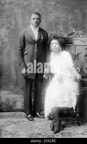 African-American newlywed couple on the day after their wedding, posing for a studio portrait, the man standing at attention and wearing a suit, the woman sitting in a chair and wearing a white dress, captioned 'Beaulah and Lloyd McIlvain', April 20, 1923. Stock Photo