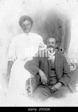 African American couple posing for a portrait photograph, man seated on a bench and wearing a suit and tie, woman wearing white dress standing behind him, Atlantic City, New Jersey, 1932. Stock Photo