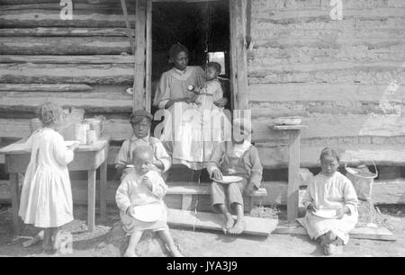 Family of african-american sharecroppers, the mother sitting in the door of a log cabin and holding young baby, several other children sitting and standing on the broken steps of the cabin and eating a meal, using their hands to eat food from bowls, each child wearing a simple dress or overalls, one girl standing at a table to the left and helping herself to food, dusty ground underneath, all children barefoot, 1910. Stock Photo