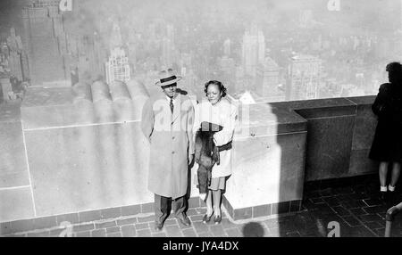 African-American couple, a man and a woman, standing together near a railing at the top of the Empire State Building, on an observation deck, viewed from above, the skyline of New York visible in the background, the man wearing a trench coat and formal hat, the woman wearing a shirt and holding a fur coat, 1946. Stock Photo