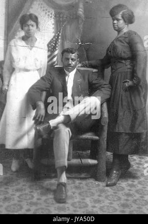 Portrait of an African American family, the father sitting in a chair with the mother standing resting her hand on the chair, daughter standing to the left of the parents, or painted backdrop featuring an American flag, 1910. Stock Photo