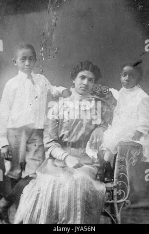 African-American mother in formal dress posing for a studio portrait, sitting in a chair, her two children standing at her sides, a boy and girl, August 20, 1910. Stock Photo