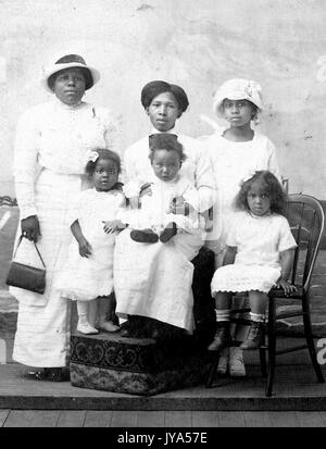 African-American family, a mother standing with her five daughters and posing for a studio portrait, all wearing white, the mother holding a purse and wearing a hat, her daughters of varying ages sitting on chairs, 1925. Stock Photo