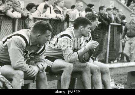 Candid photograph of three male Johns Hopkins University lacrosse players seated on the sideline bench, dressed in uniform, heads turned to the side watching the action of the game, with a crowd of onlookers standing behind a fence in the background, 1960. Stock Photo