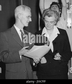 American television host Lynn Poole and television producer Mary Adams are standing and talking on the set of the television show The Johns Hopkins Science Review, Poole is wearing a business suit and is holding a packet of paper that Mary is looking over, Mary is holding a cigarette, Allan Holmes is behind Mary in a white t-shirt and is adjusting equipment, 1950. Stock Photo