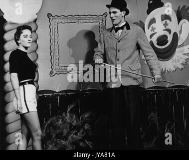 Two actors are on set filming for The Johns Hopkins Science Review, actor Lynn Rosenthal is on the left and is wearing light colored shorts and a black short sleeved shirt, actor Edmond Levy is to the right and he is wearing a checkered suit jacket with dark pants, he is holding a skinny cane, behind the two actors is painted clown's face on a wall, 1955. Stock Photo