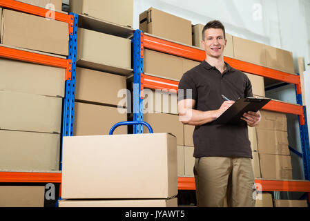 Male Worker With Cardboard Boxes Writing On Clipboard In Warehouse Stock Photo