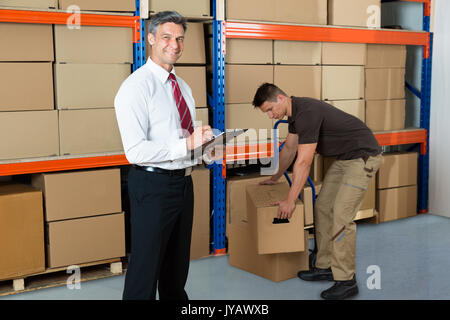 Happy Manager Writing On Clipboard While Worker Carrying Box In Warehouse Stock Photo