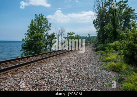 Railroad tracks on the edge of Lake Champlain with trees and crushed stone and a blue sky with a few clouds Stock Photo