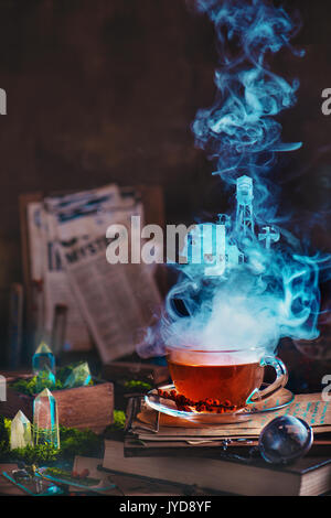 Witch kitchen. Steaming glass cup of tea with herbs and berries in a magical still life with moss and crystals. Newspaper clips on tablet on a backgro Stock Photo