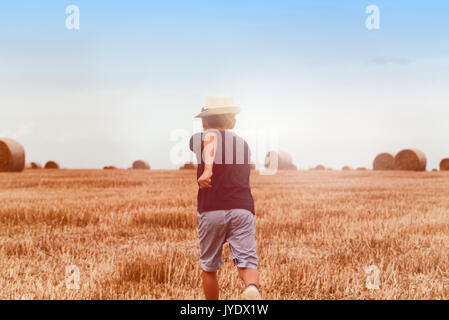 Young cute country boy in hat running happily through wheat field near hay stack or bale. Active outdoors leisure with children on warm summer day. Stock Photo