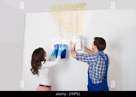 Young Woman And Repairman With Bucket Collecting Water From Damaged Ceiling Stock Photo