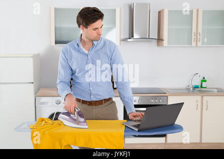 Young Man Typing On Laptop While Ironing Cloth In Kitchen Stock Photo