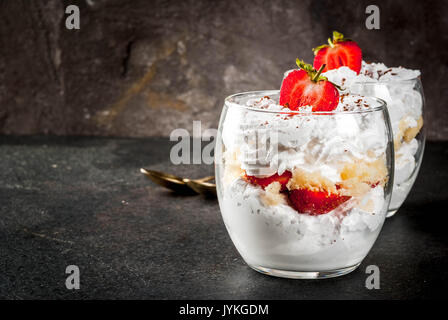Layered dessert parfait in a glass with strawberries, sponge cake and whipped cream. On a black stone table. Copy space Stock Photo