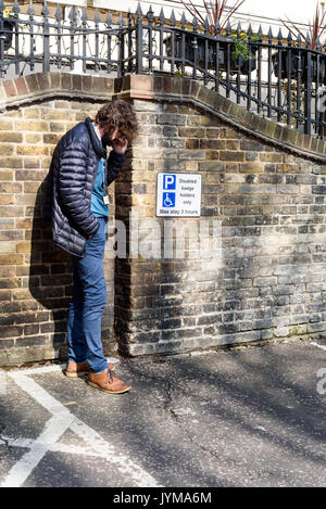 Man wearing casual clothing with hands in pocket talks on a mobile phone next to a disabled parking sign in daylight Stock Photo