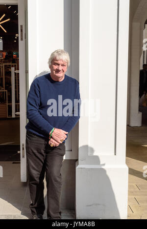 White haired man in casual clothing with hands held together leans against a white painted wall waiting utside a shop Stock Photo