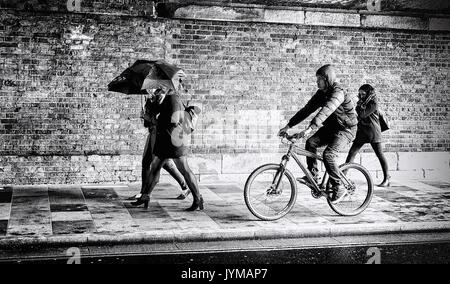 Two women with umbrellas and a man on a bike traveling through an old bridge along a pedestrian footpath Stock Photo