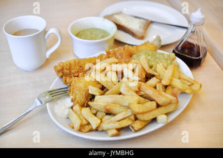 A plate of traditional British fish and chips, with mushy peas, bread and butter, vinegar, tartare sauce, and a cup of tea. Stock Photo