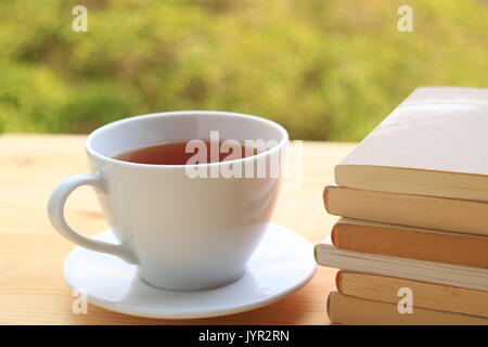 Cup of Hot Tea with Pile of Books on Wooden Table with Blurred Background of Plants in the Garden Stock Photo