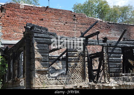 Charry corner of a burnt house with ruined wooden framework against a red brick wall with green trees and the blue sky. Stock Photo