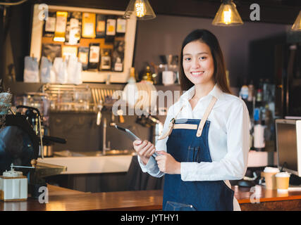 Asian female barista wear jean apron hold tablet computer coffee menu at counter bar with smile face,cafe service concept.