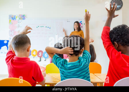 Preschool kid raise arm up to answer teacher question on whiteboard in classroom,Kindergarten education concept. Stock Photo