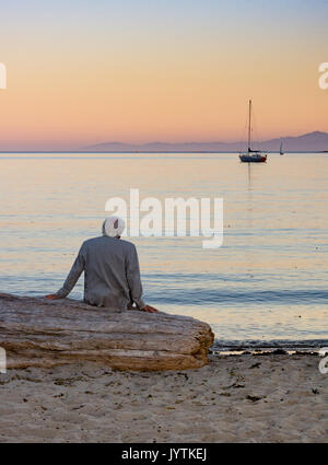 A gray haired man in a beige jacket seated on driftwood looking at the sailboats on the ocean at sunset. Stock Photo