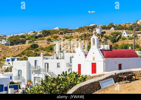 A view of white church with red roof and flying airplane on blue sky over Mykonos town, Cyclades islands, Greece Stock Photo