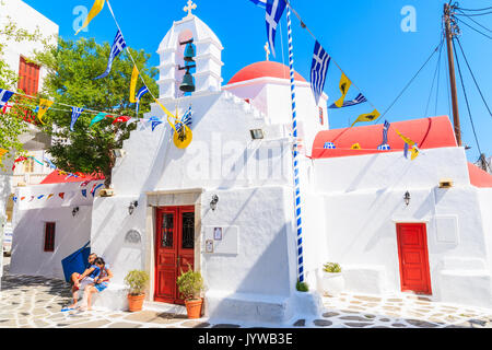 MYKONOS ISLAND, GREECE - MAY 17, 2016: Couple of young tourists resting on small square with church building on whitewashed street in beautiful Mykono Stock Photo