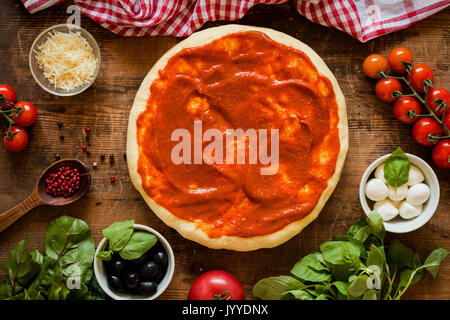 Prepared pizza dough with tomato sauce and ingredients on rustic wooden background. Cherry tomatoes, basil, mozzarella cheese, parmezan, black olives, Stock Photo