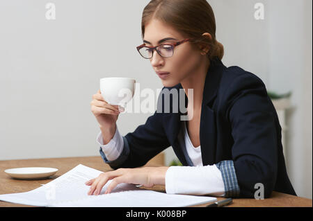 Pretty business woman reading data documents sitting at the table in office and drinking coffee Stock Photo