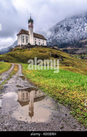 The Church of Wassen is reflected in a puddle after a winter snowfall. Andermatt. Canton of Uri. Switzerland. Europe Stock Photo