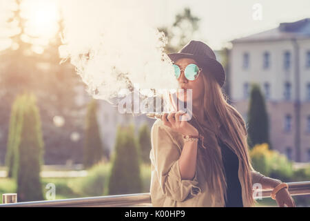 Pretty young hipster woman in black hat vape ecig, vaping device at the sunset. Toned image. Stock Photo