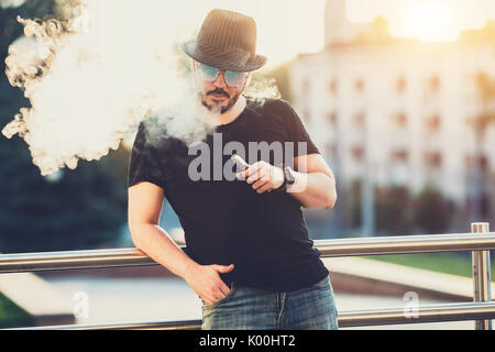 Modern happy young man in hat with a beard fun Vaporizers, In the background, the evening sunset over the city. toned image. Stock Photo