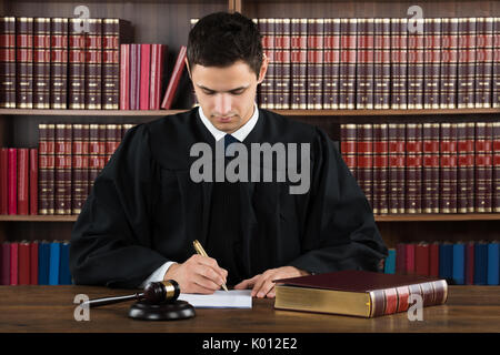 Young male judge making legal documents at desk against bookshelf in courtroom Stock Photo