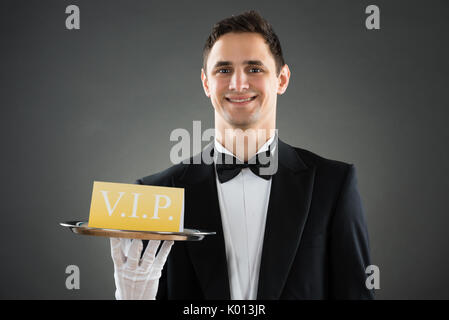 Portrait of happy young waiter holding tray with VIP sign against gray background Stock Photo