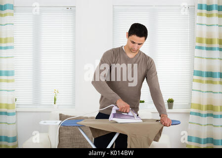 Young man ironing tshirt on board against windows at home Stock Photo