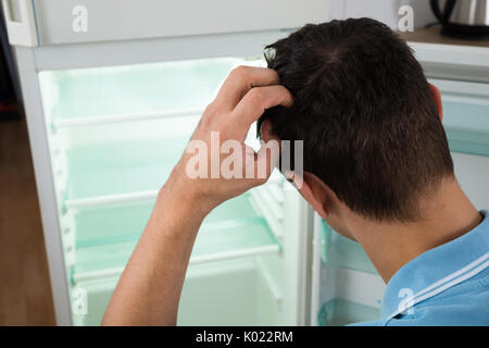 Rear view of confused man scratching head while looking at empty refrigerator at home Stock Photo