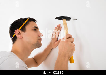 Side view of young man hammering nail on white wall at home Stock Photo