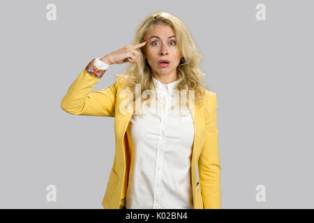 Shocked girl holding finger on temple. Portrait of surprised blonde business woman remembered something and holds a fingers on forehead, grey backgrou Stock Photo