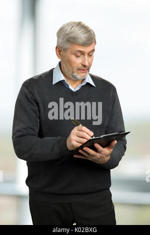 Mature man writing on clipboard. Handsome elderly man writing on black clipboard, blurred background. Stock Photo