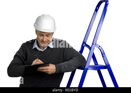 Portrait of mature building inspector. Senior engineer leaning on ladder and writing on clipboard, white background. Foreman checking new object. Stock Photo