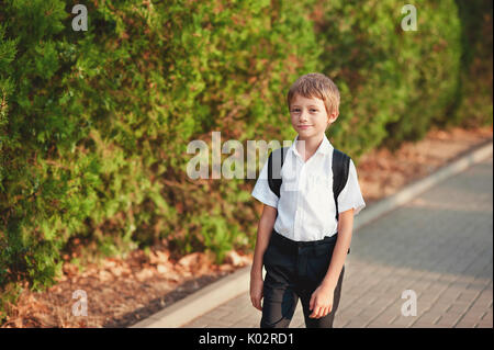 Little schoolboy comes back from school in good mood. The boy goes on a footpath. He has raised eyes to the sky and smiles. Behind shoulders at the pupil a satchel. Stock Photo