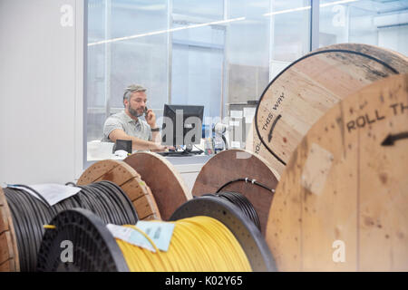 Male supervisor working at computer and talking on ell phone behind spools in fiber optics factory Stock Photo