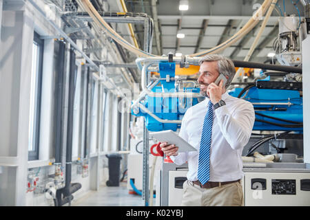 Male supervisor with digital tablet talking on cell phone next to plastic industrial molder machinery in factory Stock Photo