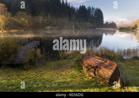 An upturned old wooden boat lying on grass on a misty morning with reflection, Loch Ard, Scotland Stock Photo