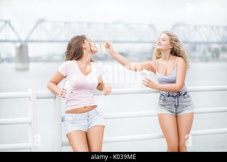 Beautiful young women, leaning on parapet, sharing a fast food lunch box, eating up noodles from Chinese take-out with chopsticks and drinking takeawa Stock Photo